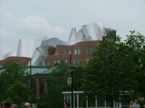 A modern building with a wavy silver roof and brick walls, surrounded by trees and people in a park setting.