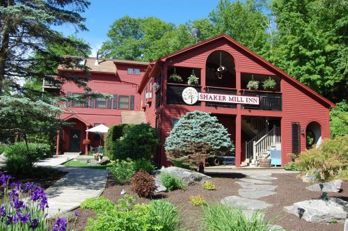 A red inn surrounded by greenery, featuring a sign that reads "Shaker Mill Inn" and colorful flowers in the foreground.