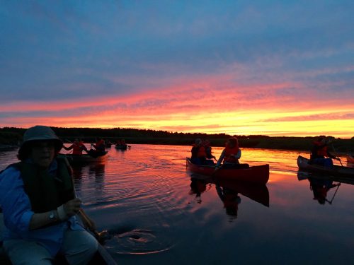 Canoers paddle on a calm river at sunset, with vibrant colors reflecting on the water.