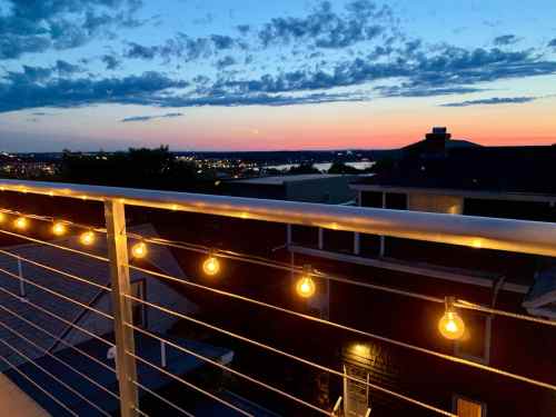 A sunset view from a balcony with string lights, overlooking a city and water in the distance.