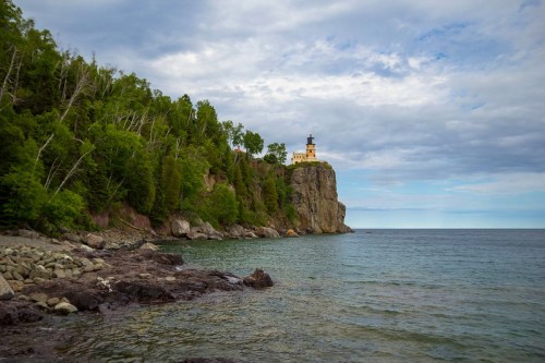 A lighthouse perched on a rocky cliff surrounded by lush greenery and calm waters under a cloudy sky.