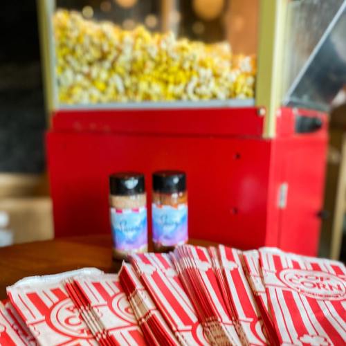 A popcorn machine with colorful popcorn, two seasoning containers, and striped popcorn bags on a table.