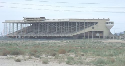 Abandoned stadium with a partially collapsed structure, surrounded by overgrown grass and sparse vegetation.