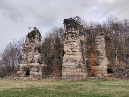 Three tall, rocky formations rise from a grassy field, surrounded by bare trees under a cloudy sky.