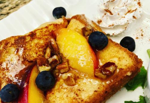 A slice of French toast topped with peaches, blueberries, pecans, and a dollop of whipped cream on a white plate.