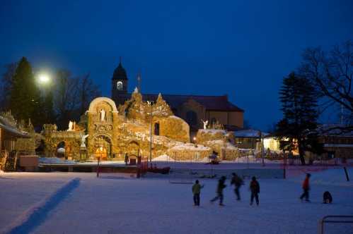 A snowy landscape at dusk with a lit ice sculpture and people skating in the foreground.