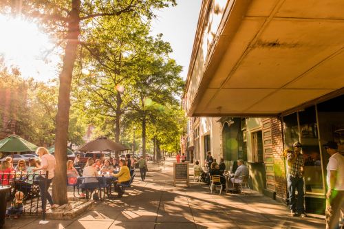 A sunny street scene with people dining outdoors at tables, surrounded by trees and shops.
