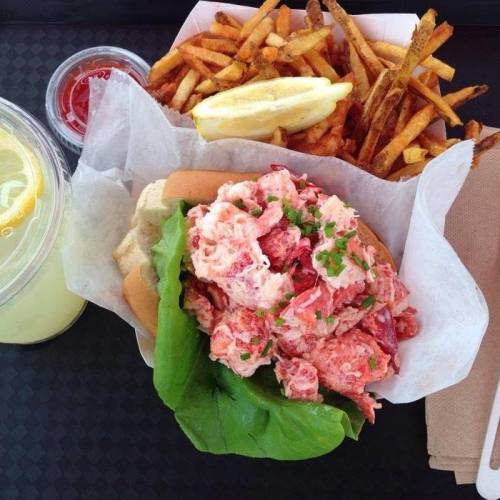 A lobster roll with lettuce, served with fries and a lemon wedge, alongside a drink.