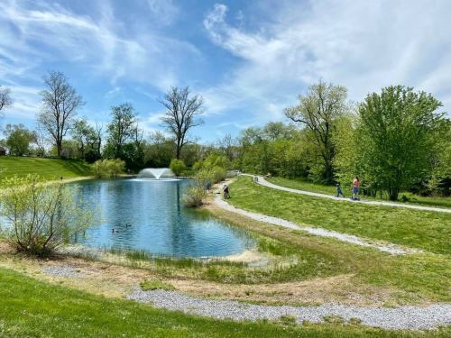 A serene park scene featuring a pond, walking path, trees, and people enjoying the outdoors on a sunny day.