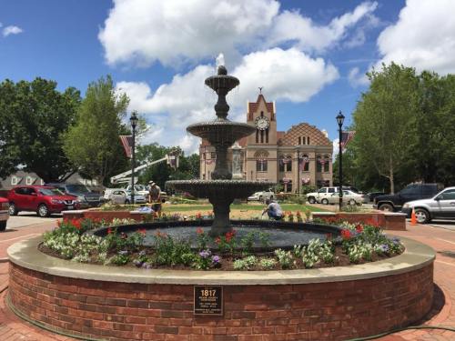 A decorative fountain surrounded by flowers in a town square, with a historic building and cloudy sky in the background.