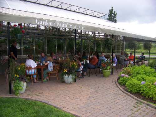 Outdoor café with patrons dining at tables surrounded by greenery and flowers under a canopy.