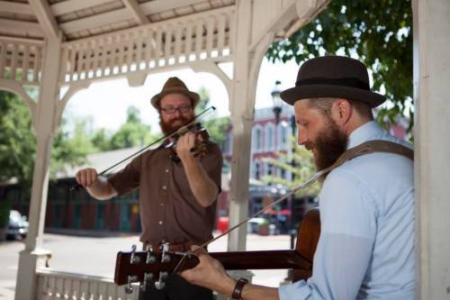 Two musicians perform in a gazebo; one plays the guitar while the other plays the violin, both wearing hats.