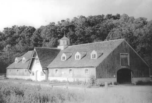 A vintage black-and-white photo of a large barn surrounded by trees, featuring a cupola and multiple windows.