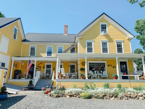 A charming yellow house with a porch, surrounded by flowers and greenery, under a clear blue sky.