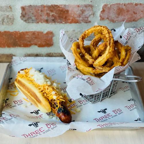 A hot dog topped with onions and cheese, served with a side of crispy onion rings in a small basket.