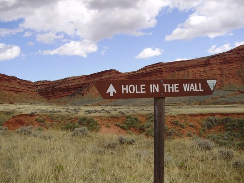A brown sign pointing to "Hole in the Wall" with red rock formations and a cloudy sky in the background.