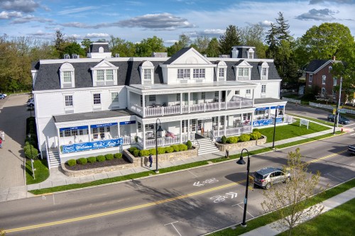 A large white building with a porch, surrounded by greenery, located on a street with parked cars and pedestrians.