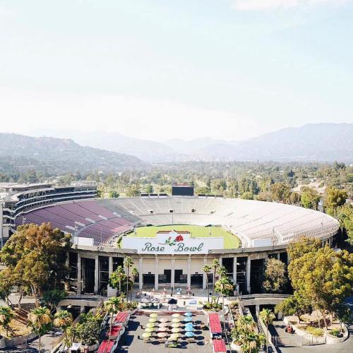 Aerial view of the Rose Bowl stadium surrounded by trees and mountains, featuring a colorful outdoor seating area.