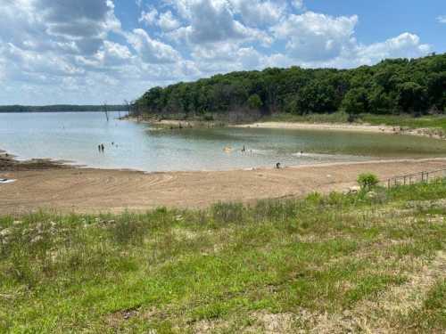 A sandy beach by a calm lake, with people swimming and trees lining the shore under a partly cloudy sky.