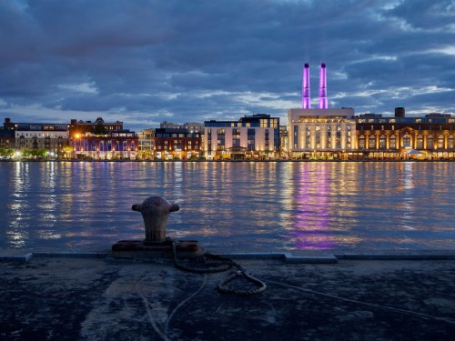 A waterfront view at dusk, featuring buildings illuminated with purple lights and a mooring post in the foreground.
