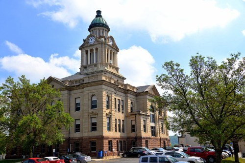 Historic courthouse with a clock tower, surrounded by trees and parked cars under a partly cloudy sky.