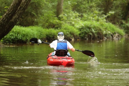 A person paddles a red kayak through a calm, green river surrounded by lush vegetation.