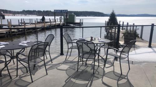 Outdoor dining area with empty tables overlooking a calm lake and a dock on a sunny day.