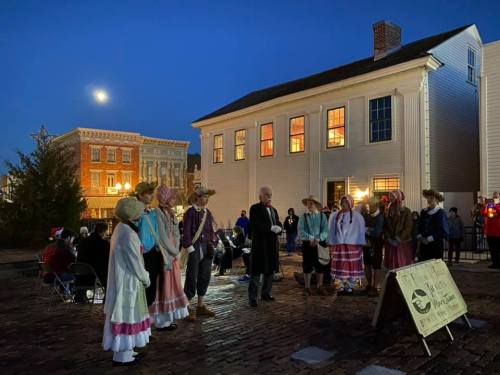 A group of performers in historical costumes gather outdoors at night, with a full moon and a historic building in the background.