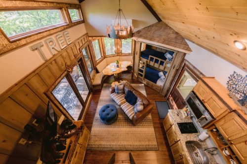 Cozy interior of a wooden cabin with a living area, kitchen, and lofted sleeping space, featuring large windows and rustic decor.