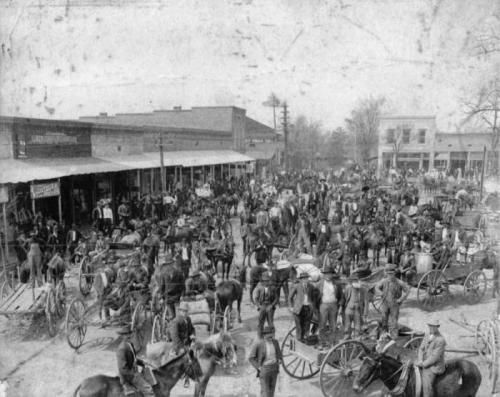 A bustling historic marketplace filled with people, horses, and wagons, set in a town with storefronts in the background.