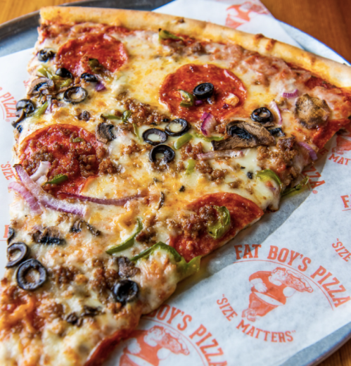 A close-up of a large slice of pizza topped with pepperoni, olives, green peppers, onions, and sausage on branded paper.