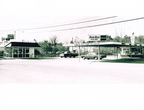 A vintage black-and-white photo of a roadside diner and parking area with cars and signage.