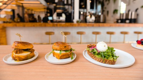 Three gourmet sandwiches on white plates, featuring cheese, greens, and a poached egg, set in a stylish café.