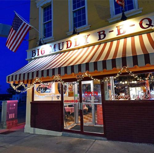 A cozy BBQ restaurant with a striped awning, bright lights, and an American flag, welcoming customers at night.