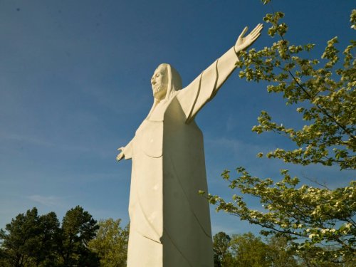 A large white statue of Jesus with outstretched arms, surrounded by trees and a clear blue sky.