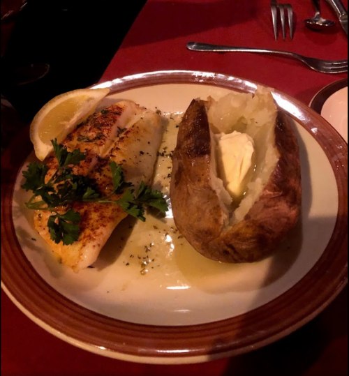 A plate with grilled fish garnished with parsley, a lemon wedge, and a baked potato with butter.