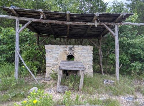 An old stone structure with a wooden roof and table, surrounded by overgrown grass and trees.