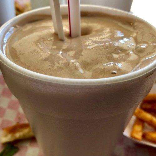 A close-up of a chocolate milkshake in a white cup with two straws, alongside a side of fries.
