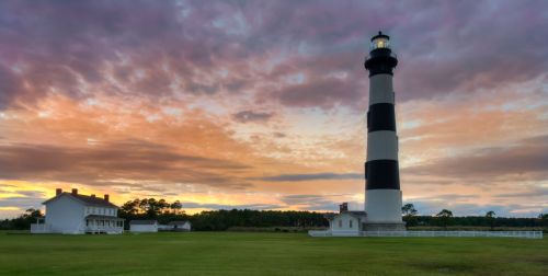 A black and white striped lighthouse stands beside a white house against a colorful sunset sky.