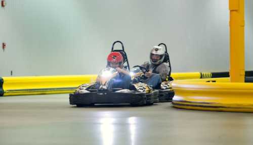 Two go-karts racing on an indoor track, with one driver in a red helmet and the other in a white helmet.