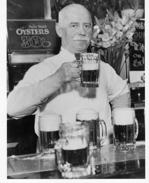 A man in a white shirt holds a large beer mug, standing behind a bar with several beer glasses in front of him.