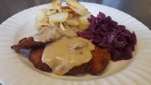 A plate with breaded meat covered in gravy, served with sautéed potatoes and purple cabbage.
