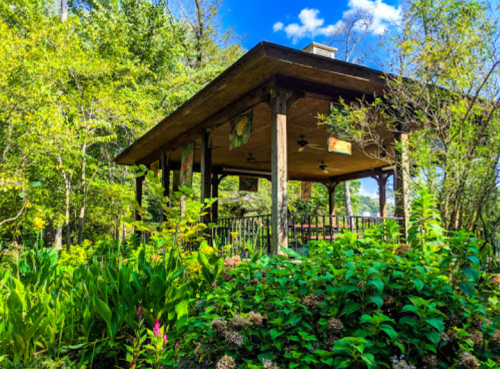 A wooden gazebo surrounded by lush greenery and colorful flowers under a bright blue sky.