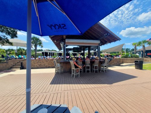 A sunny outdoor bar area with umbrellas, seating, and a pool in the background, surrounded by palm trees.