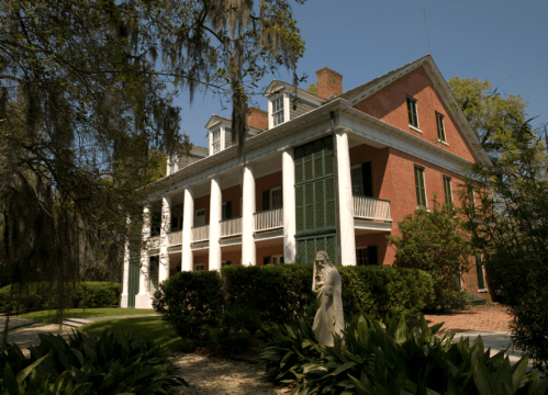 Historic brick mansion with white columns, surrounded by greenery and a statue in the foreground.