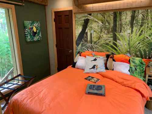 Cozy bedroom with an orange bedspread, penguin pillows, and a nature-themed wall mural. A chair and books are nearby.