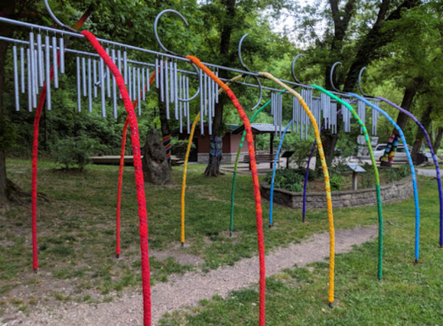 Colorful wind chimes hang from a curved metal frame in a green park, surrounded by trees and a gravel path.