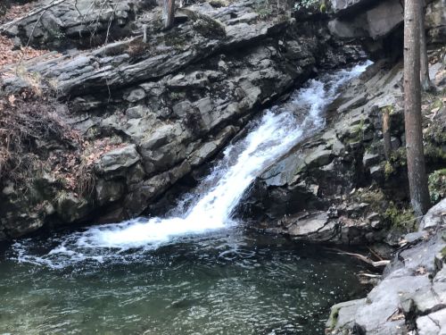 A small waterfall cascades over rocky terrain into a serene pool surrounded by trees and natural scenery.