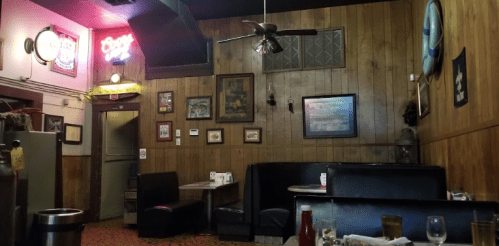 Cozy restaurant interior with wooden walls, booths, and vintage decor, featuring a ceiling fan and neon signs.