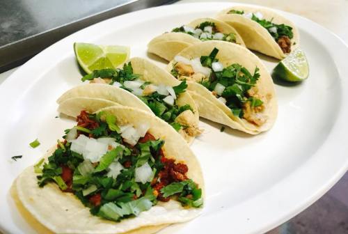 A plate of tacos with various fillings, topped with onions and cilantro, served with lime wedges.
